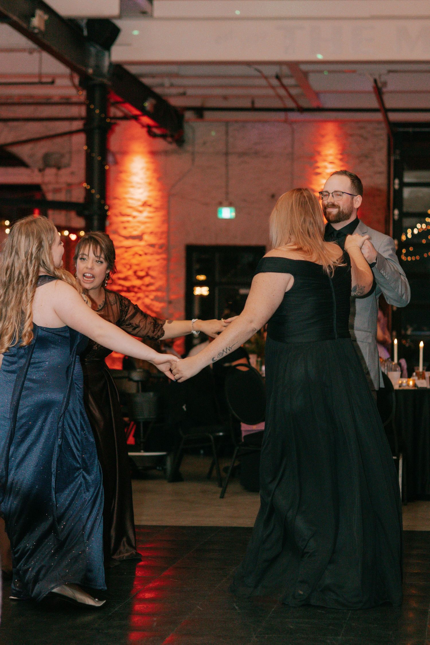 Wedding guests dancing in formal attire under ambient red lighting at an indoor venue.