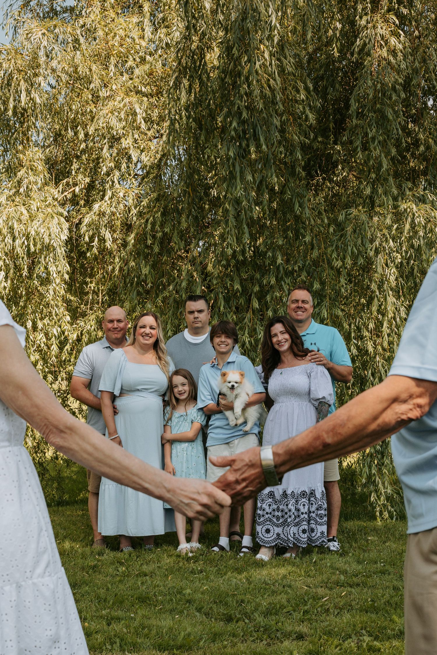Family stands in a line holding hands against backdrop of willow trees.