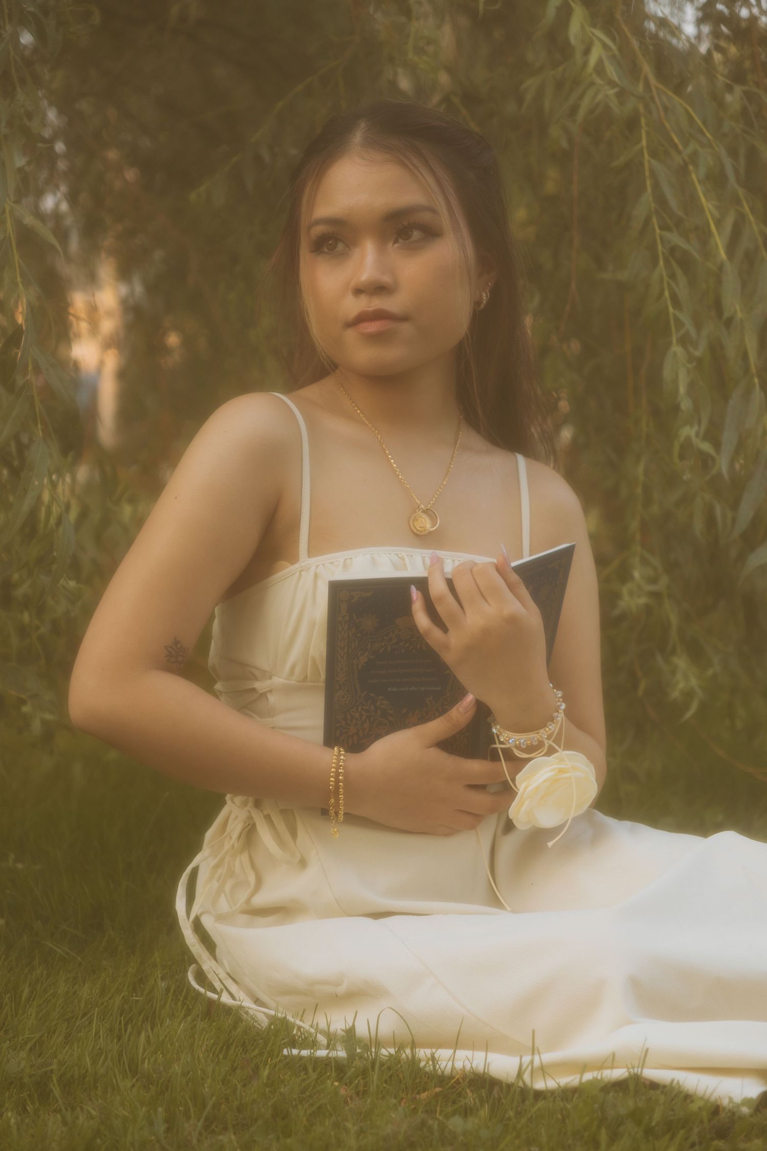 A romantic outdoor photo of someone holding a book while wearing a flowing white dress under willow branches.
