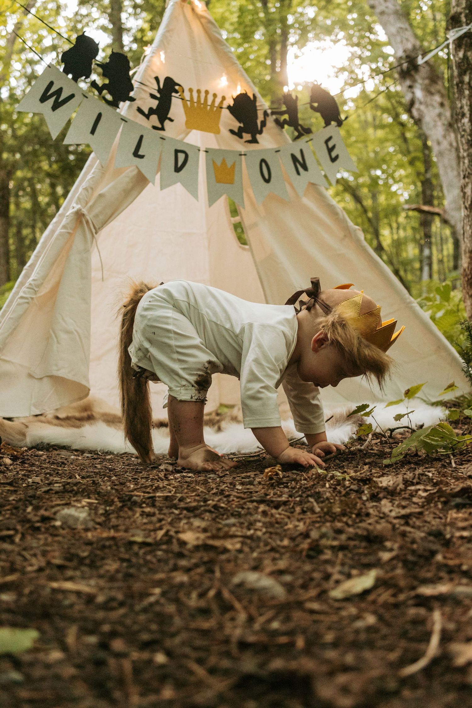 Child playing near a teepee tent with 'Wild One' banner in a forest setting.