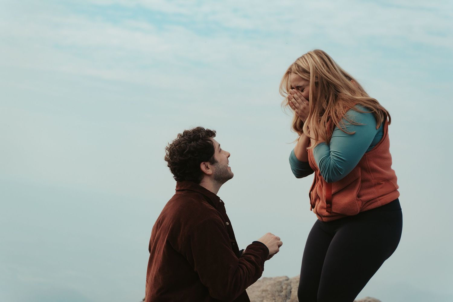 Surprise engagement mountaintop photo session with a man in red down on one knee and the skyline in the background