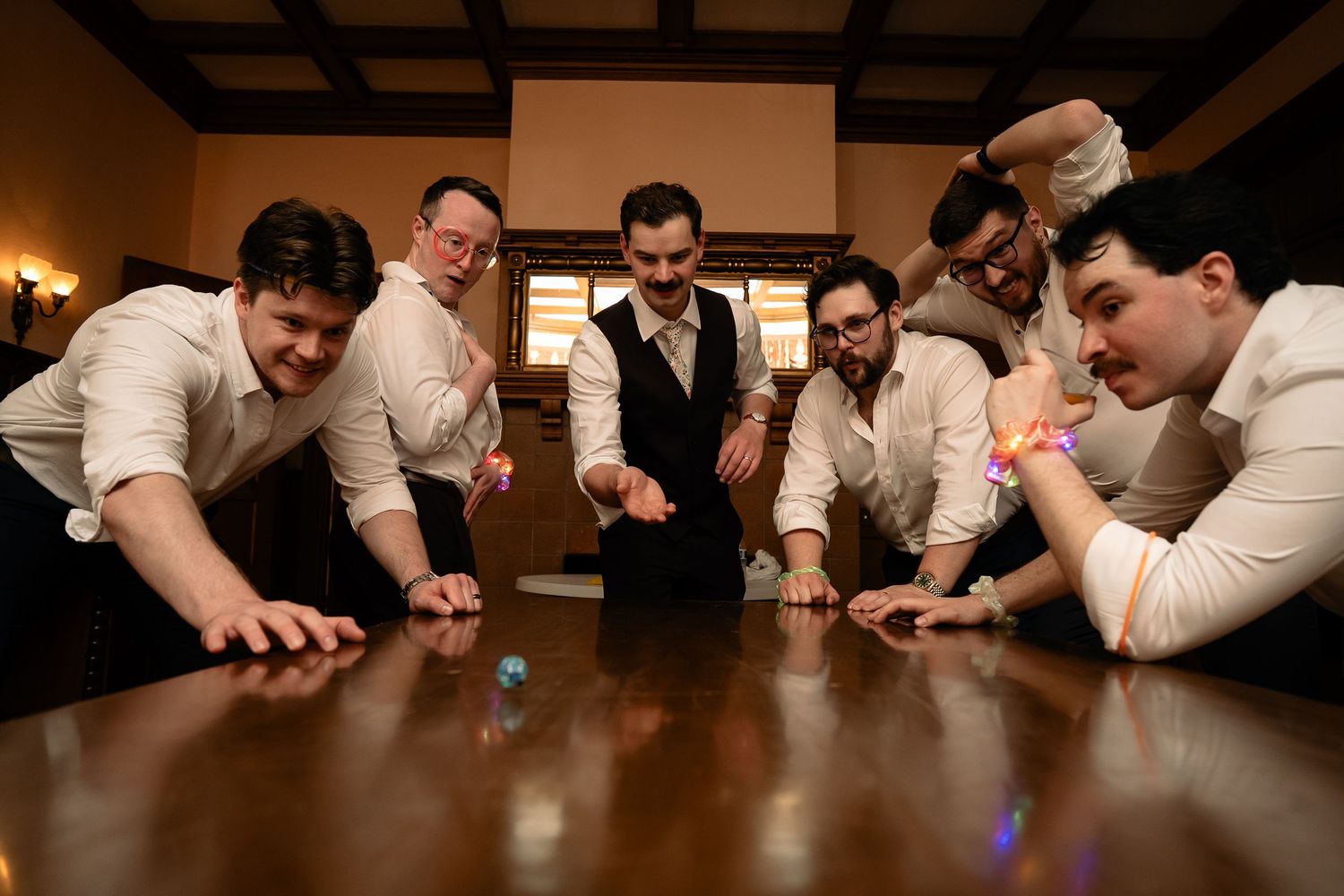 Groomsmen are held in suspense around a reflective dark wood table as a groom rolls a 20-sided die used in their Dungeons & Dragons campaign