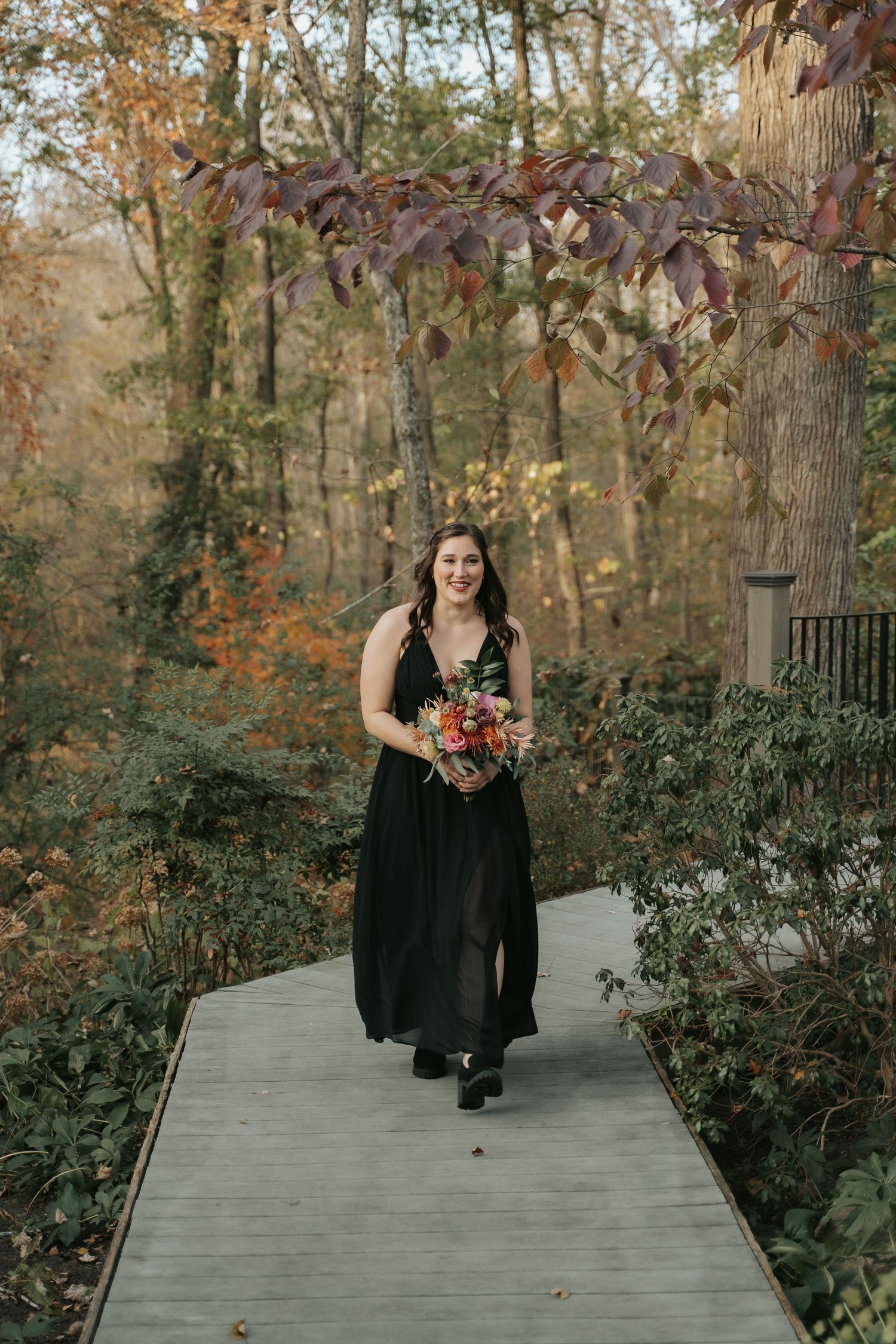 A bridesmaid in a dark dress walks with a colorful bouquet  under a fall canopy during a wedding ceremony at The Mill at Fine Creek