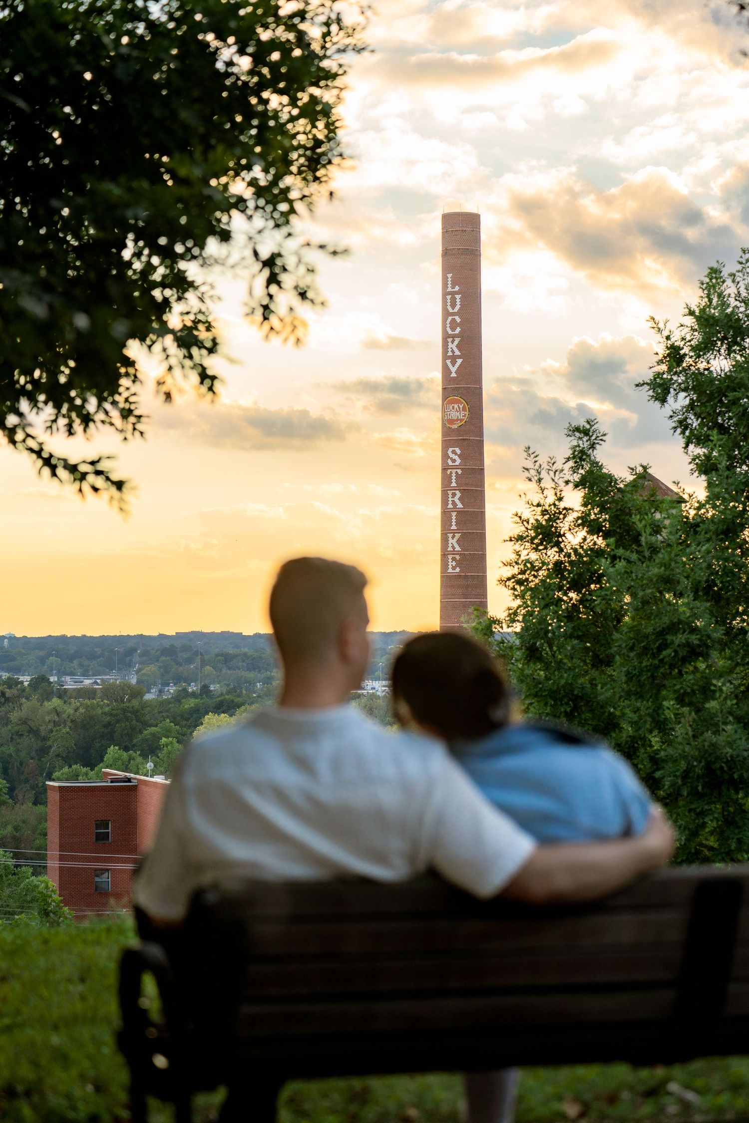 Couple hugs sitting on bench facing sunset away from camera in RVA engagement photo session in Libby Hill Park