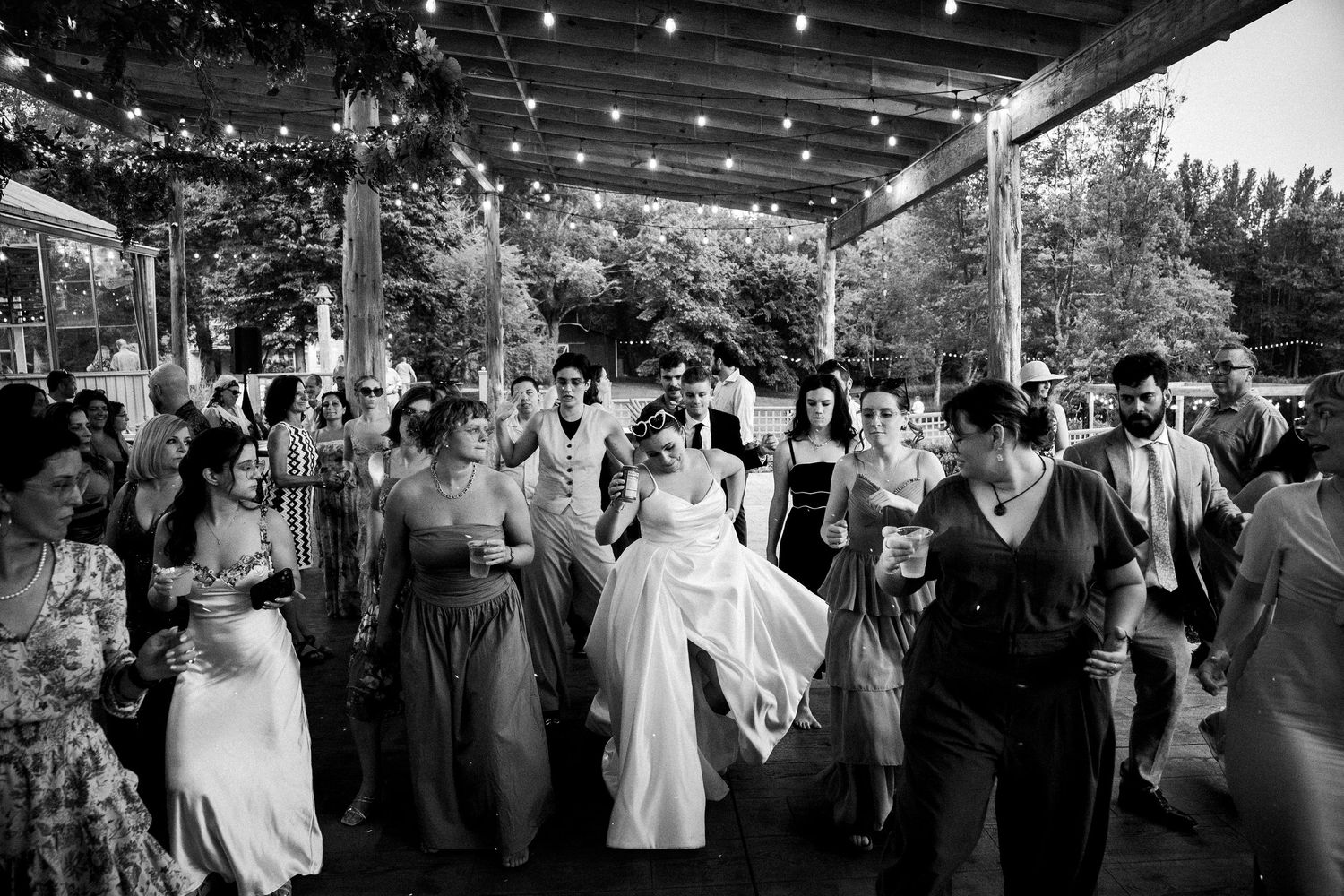 Bride in dress leads line dance of wedding guests under the covered dancefloor in documentary photograph of Avonlea Farms