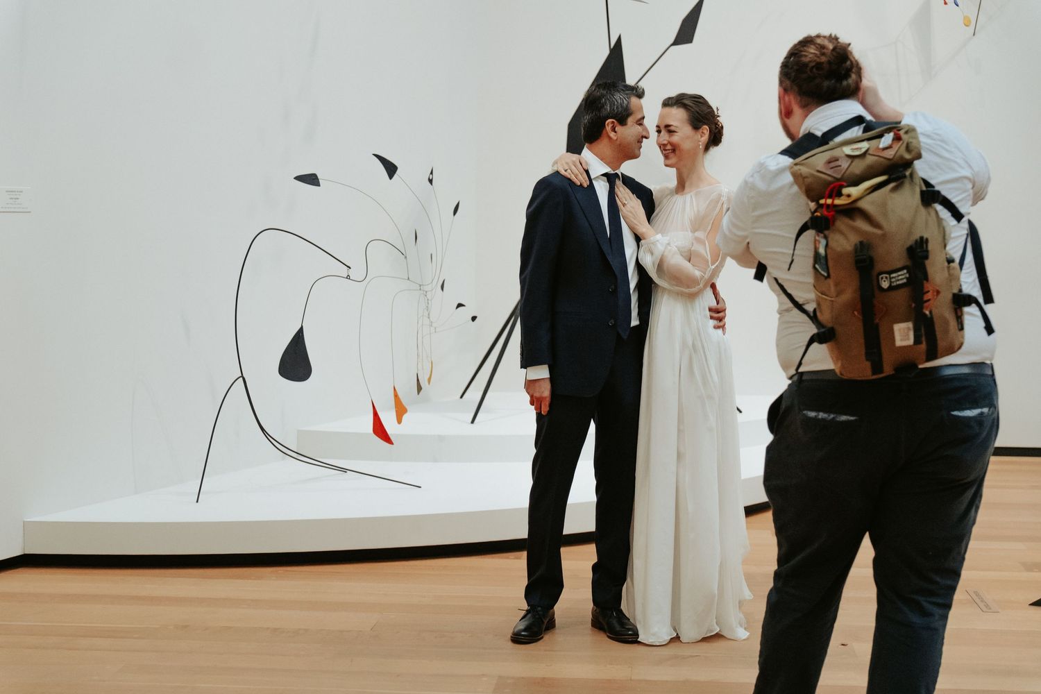 A bride in groom pose in front of a modern sculpture while an out-of-focus photographer stands in the foreground to get their portrait