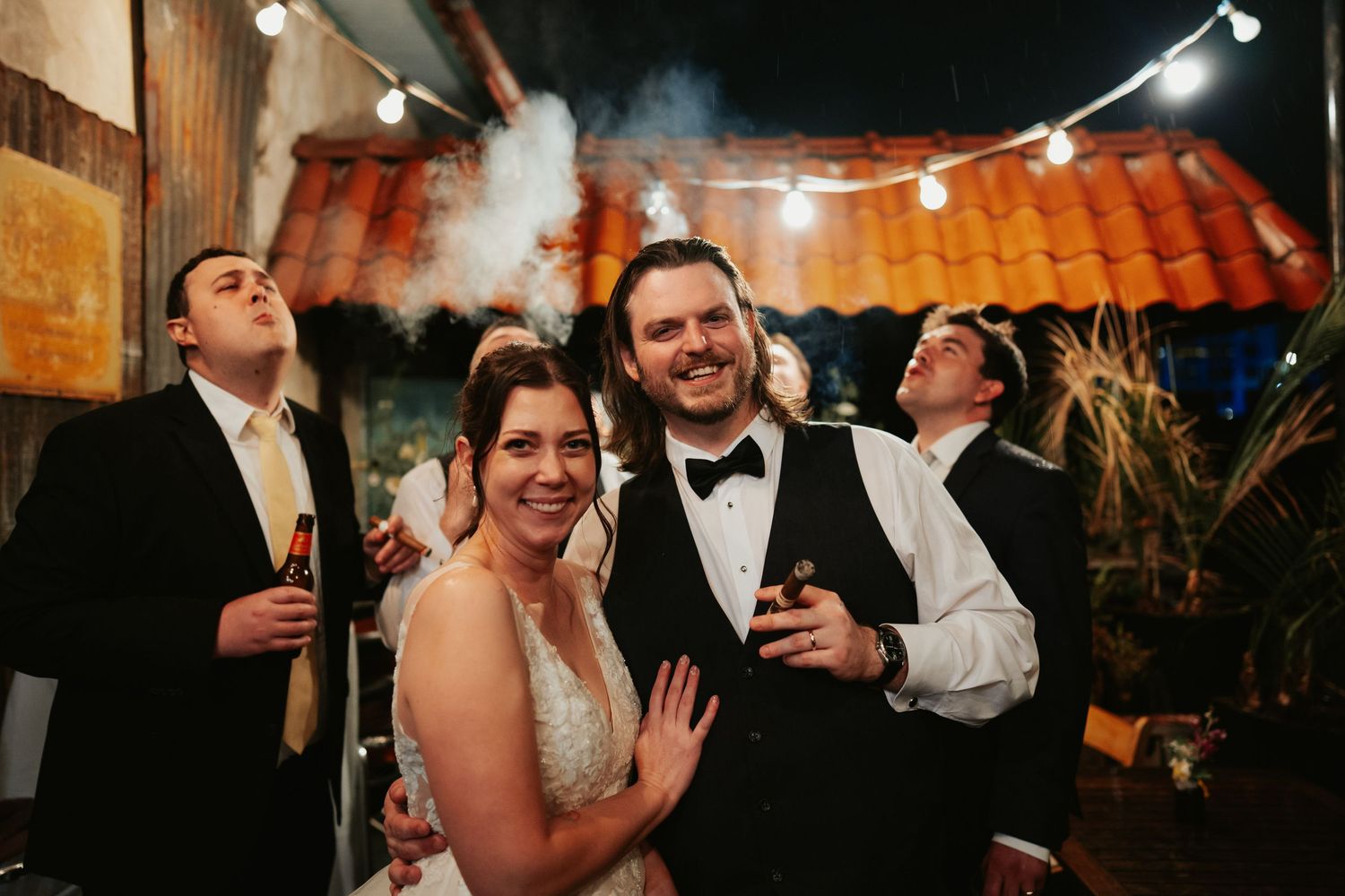 Richmond Virginia couple in wedding dress and black tuxedo smile as their groomsmen blow smoke clouds on a rooftop lit by market lights.