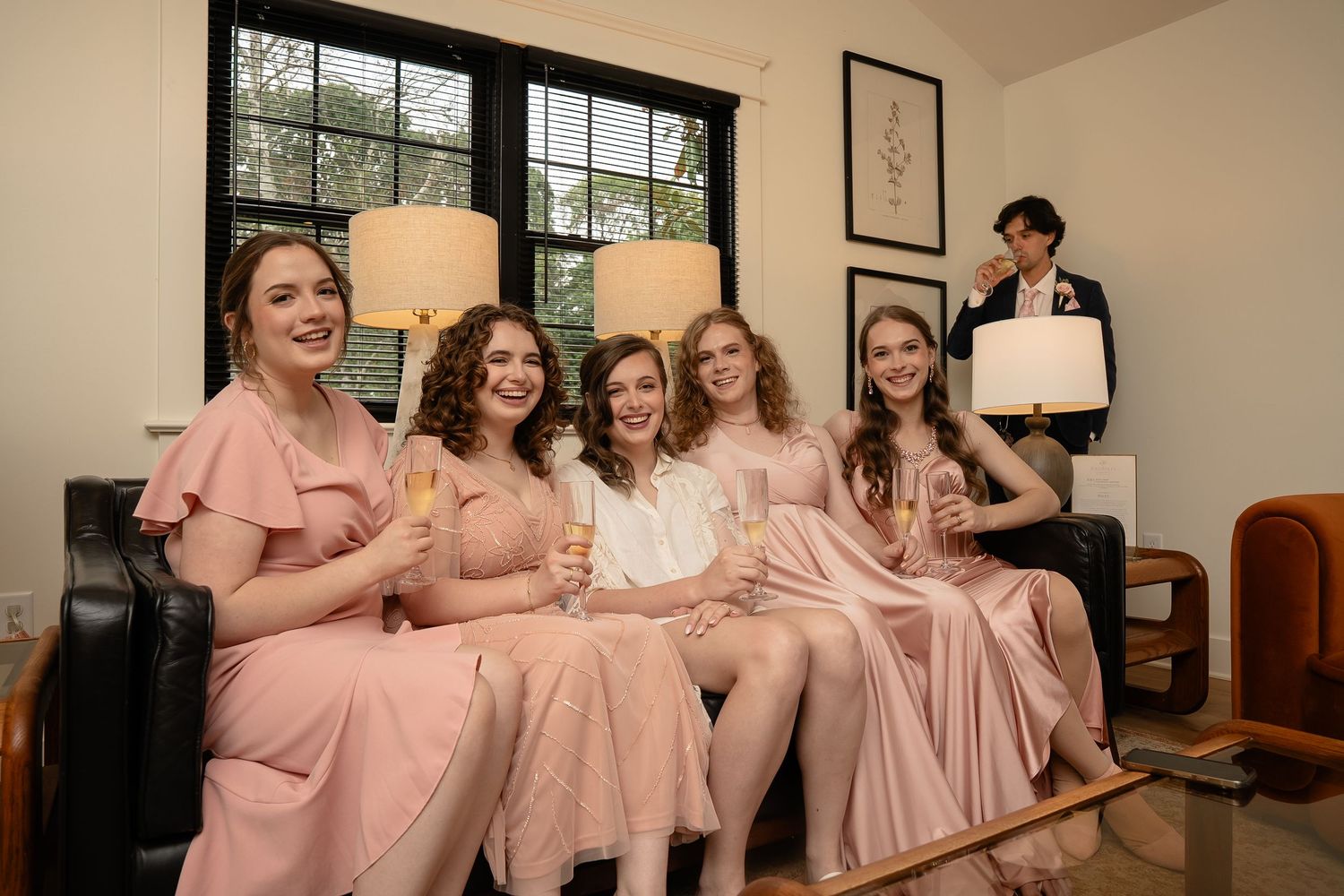 Bridesmaids pose with smiles on a couch with the bride while a bridesman pouts in the back corner at a queer wedding at The Market at Grelen