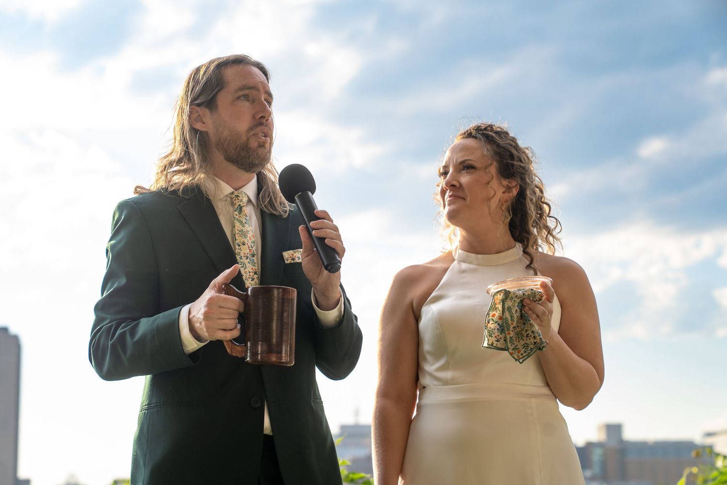 Bride in white sleeveless dress and groom in green suit with floral tie welcome their guests to the wedding against backdrop of dramatic sky