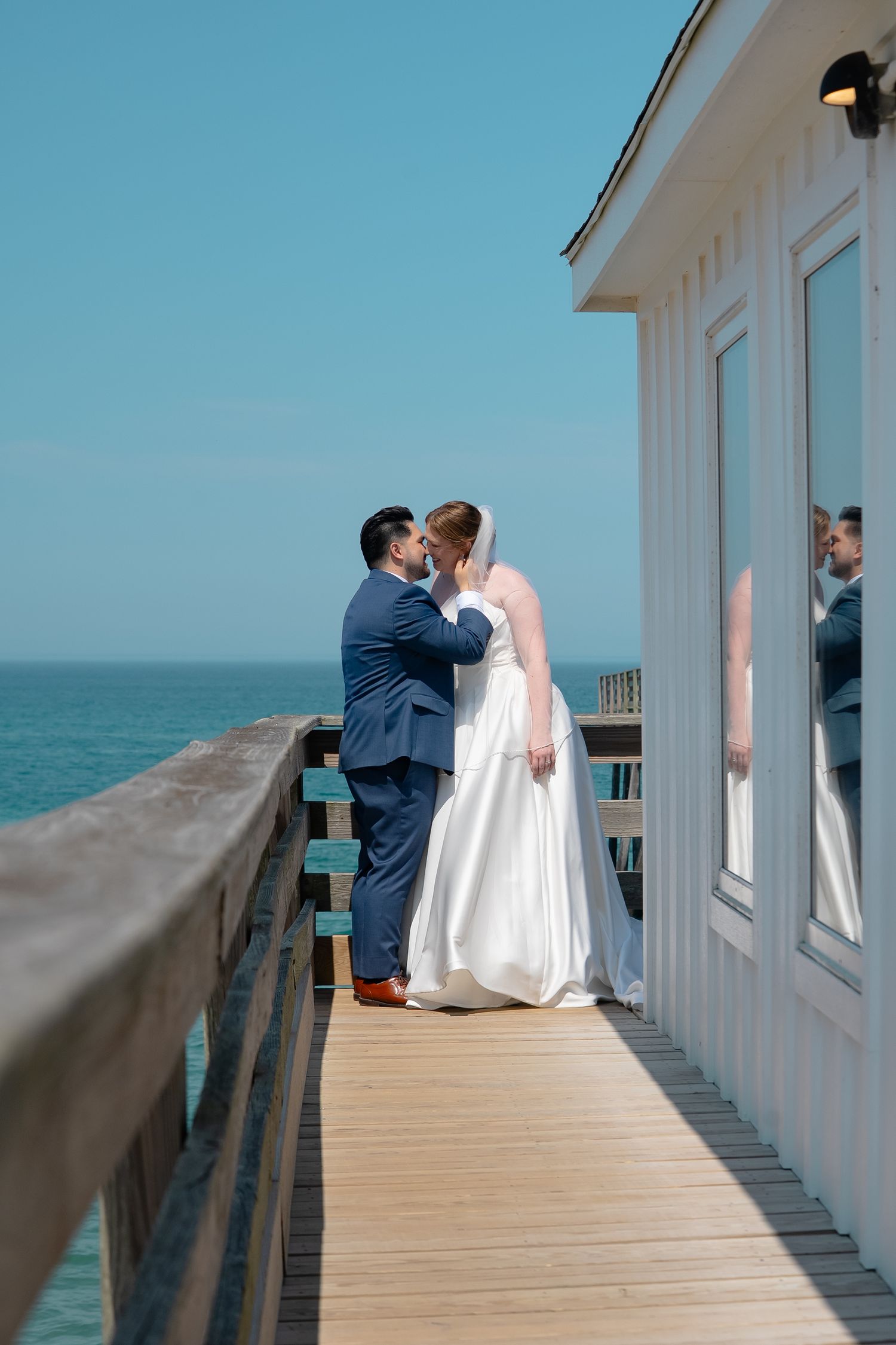 A bride and groom kiss at the end of a deck overlooking the ocean while a row of windows shows their reflection