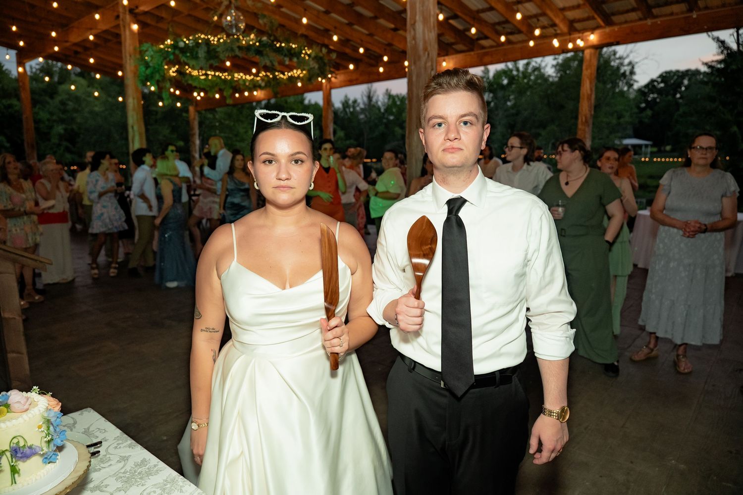 Bride and groom stand holding cake slicing utensils as if they are the couple from the painting American Gothic
