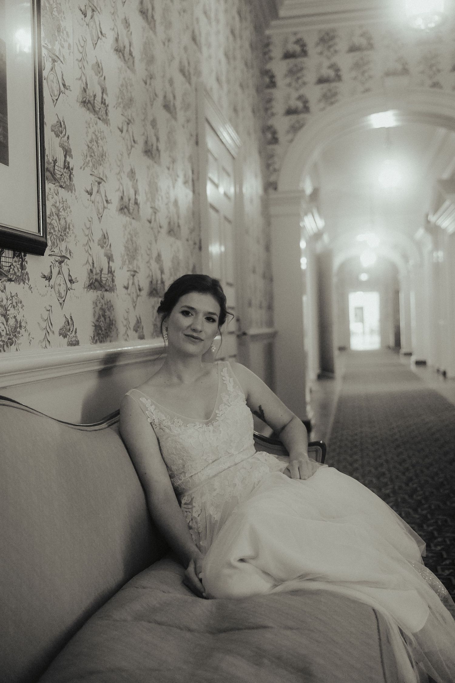 Vintage black and white aesthetic wedding photo with bride sitting on couch in historic wall-papered venue facing camera