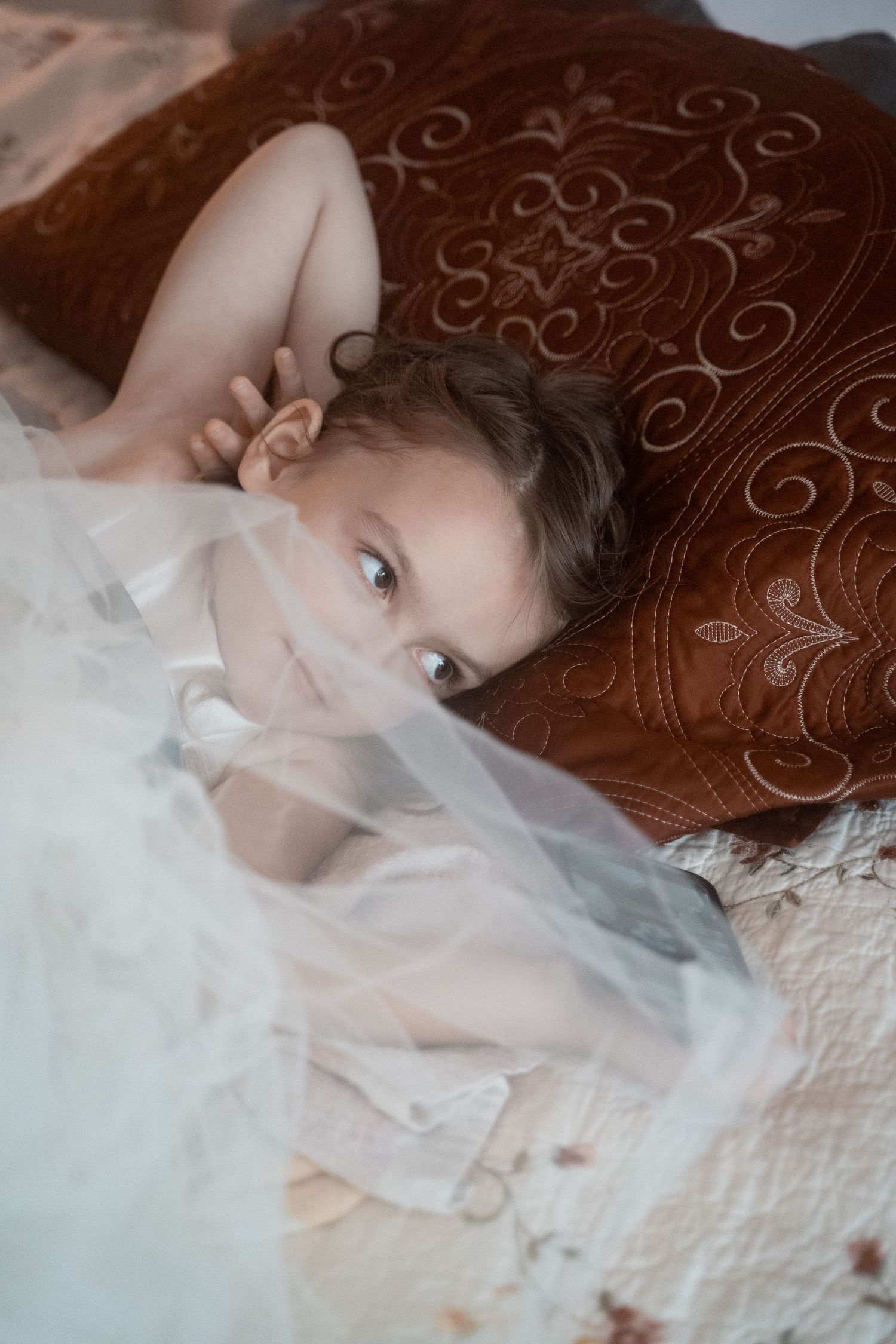 Bride's youngest sister lays on her bed under a veil while watching the Bride get ready for the day