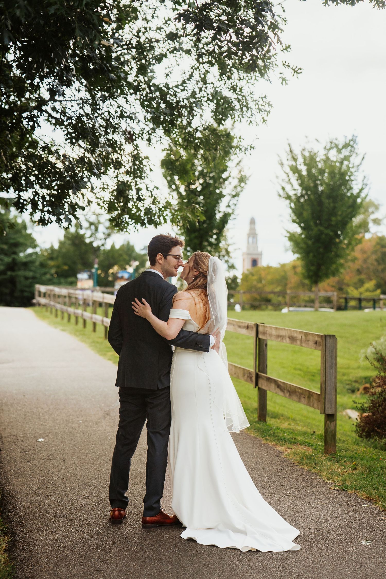 Bride and groom with backs to camera and countryside aesthetic background, bride wears an off-shoulder dress with a train and veil