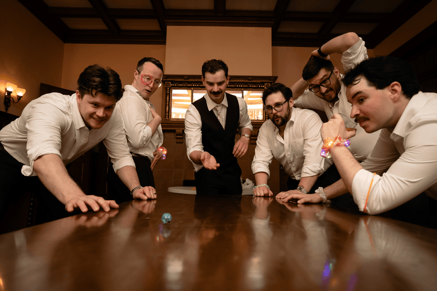 A groom in shirt and vest is surrounded by his dungeon and dragons groomsmen as he rolls a nat 20 and celebrates by jumping around the room.