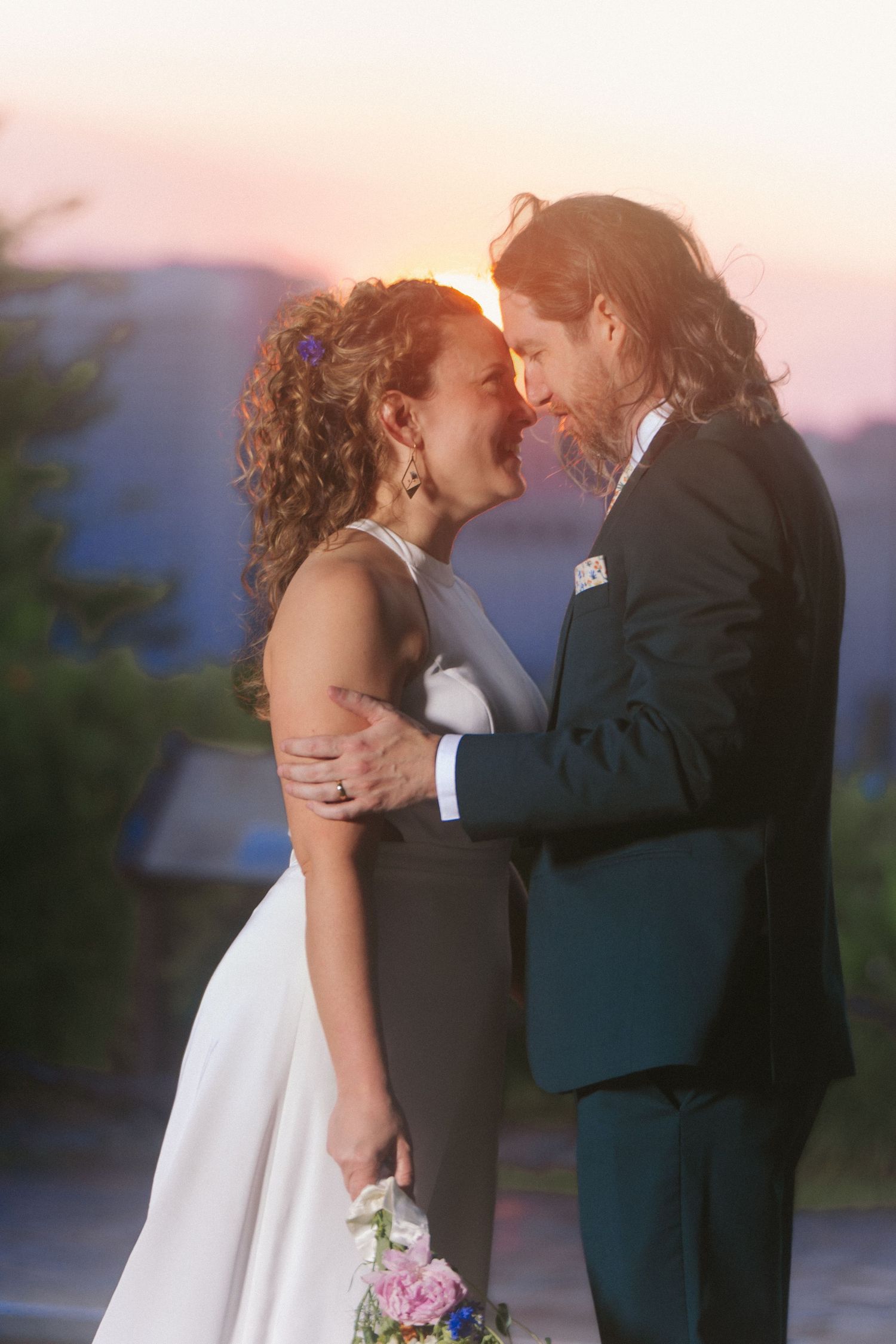 Bride and groom stand in front of sunset in classic summer wedding photo with bride in modern sleeveless dress