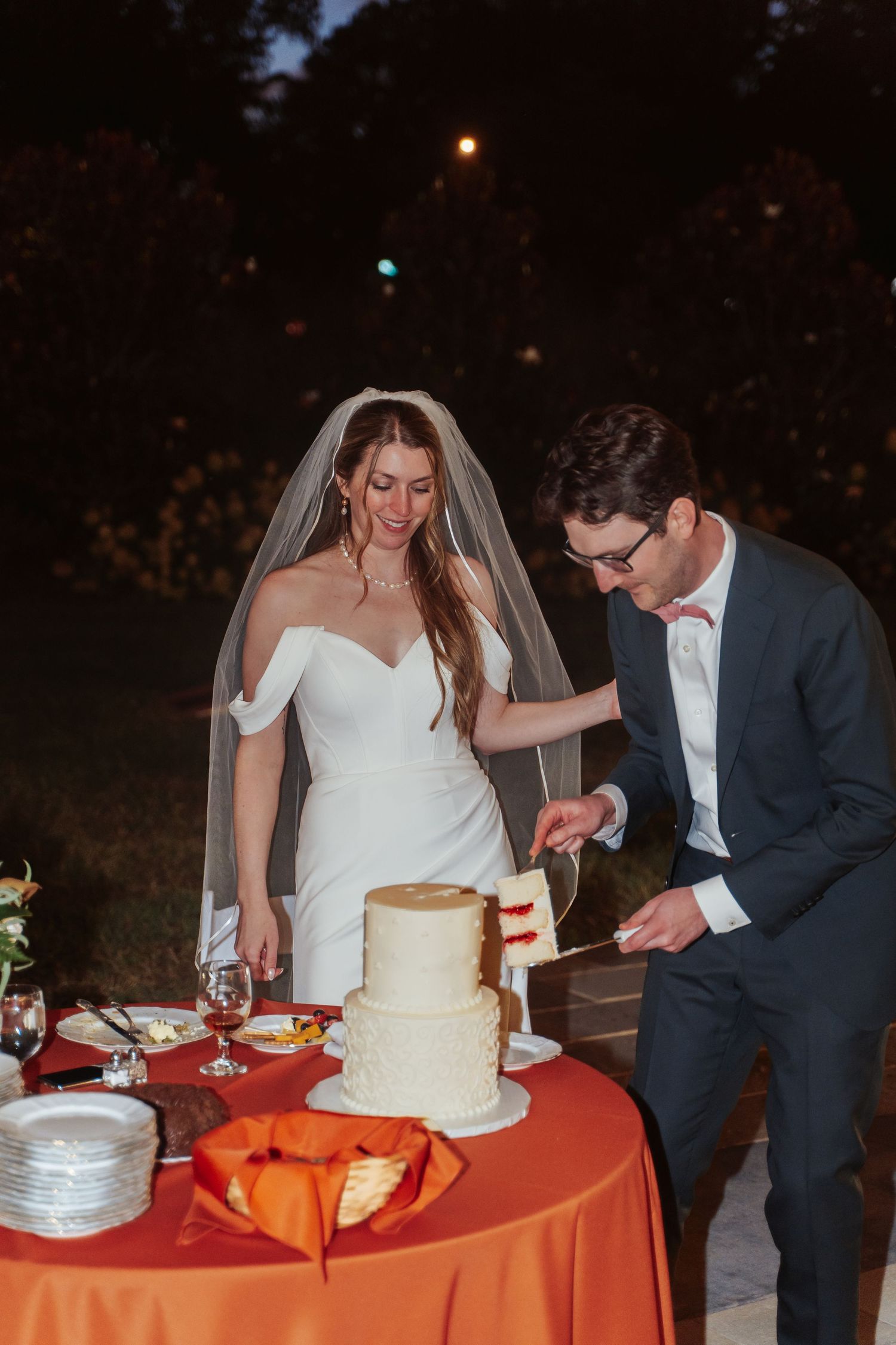 Richmond Virginia bride in a long veil watches her groom slice their wedding cake in the patio of Maymont Gardens