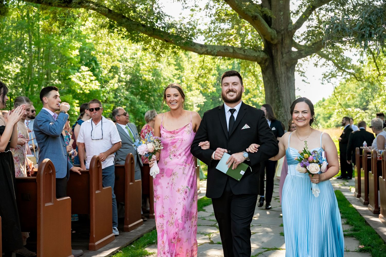 A groomsmen and two bridesmaids beam smiles as they recess out of a tree-covered ceremony at Avonlea Farms outside Richmond Virginia