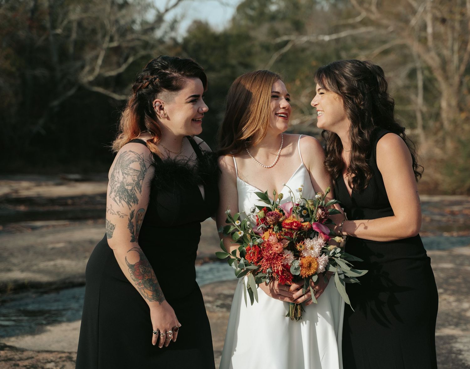Timeless bride laughs with her sisters in black bridesmaid dress on the rocks of Mill at Fine Creek for winter wedding outside Richmond