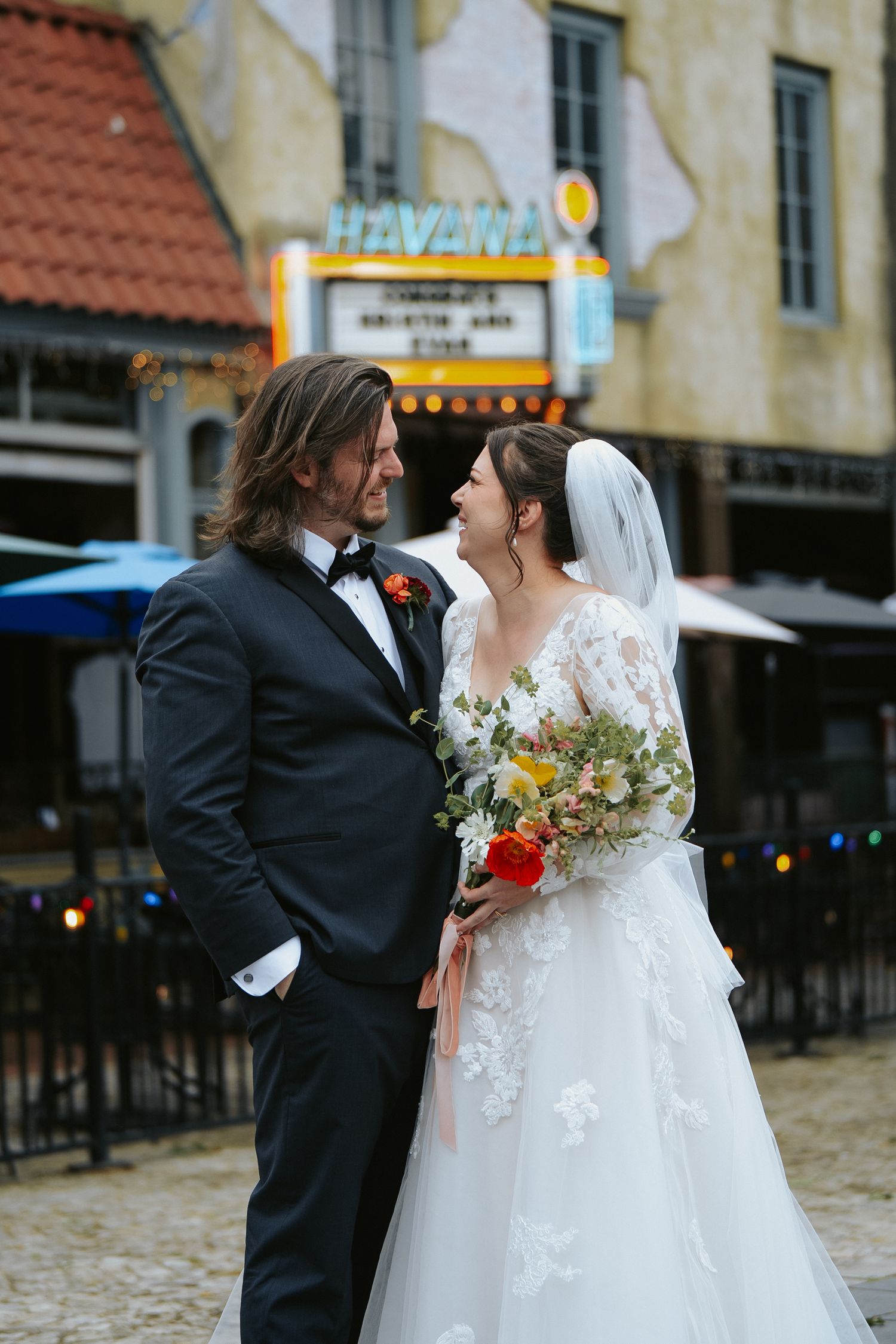 Retro vintage wedding aesthetic with bride in classic lace veil and bouquet and groom in black tux in front of marquee, street-style shoot