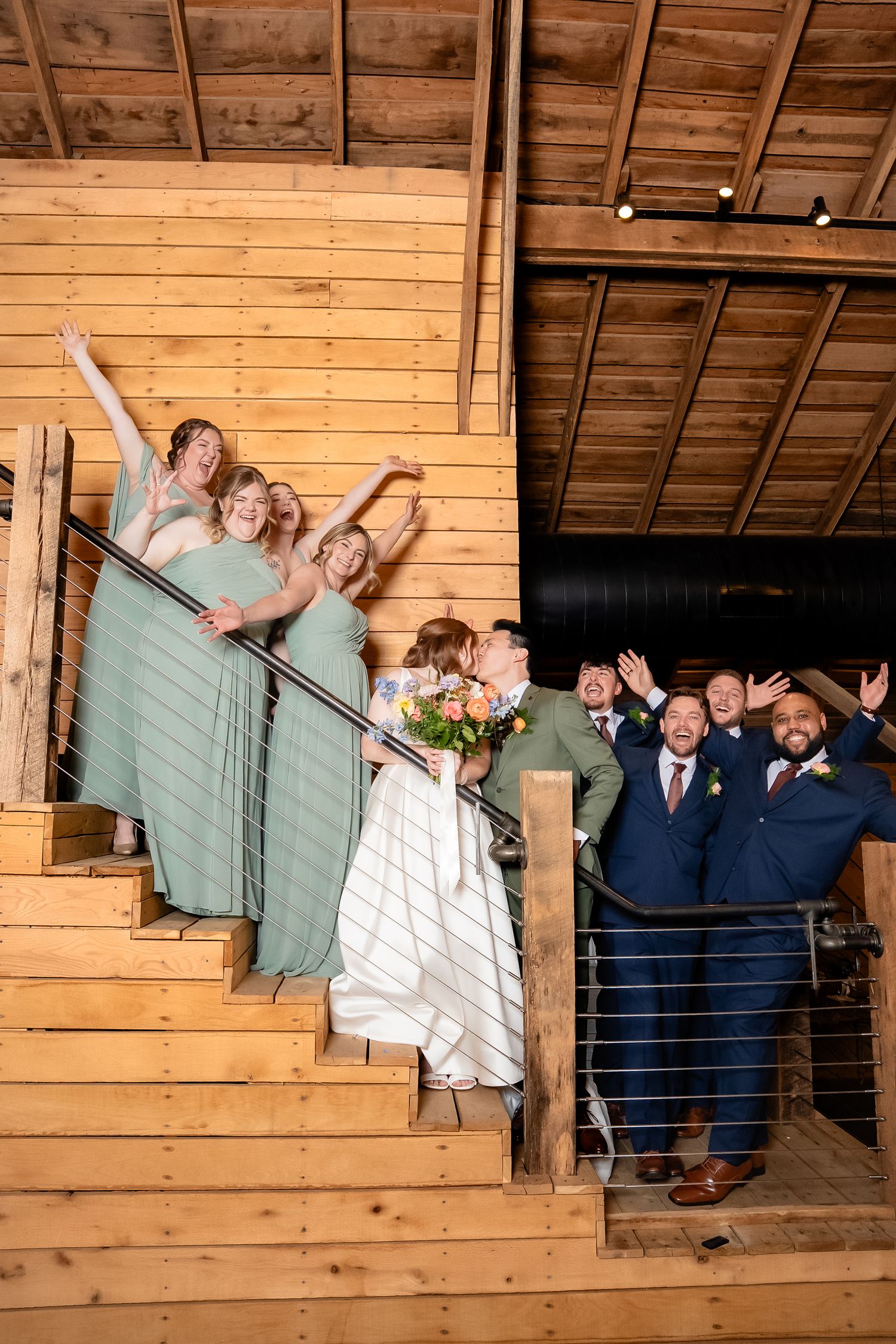 The wedding party celebrates on the stairs as the nearlyweds share a kiss between them at a venue in the Blue Ridge Mountains of Virginia