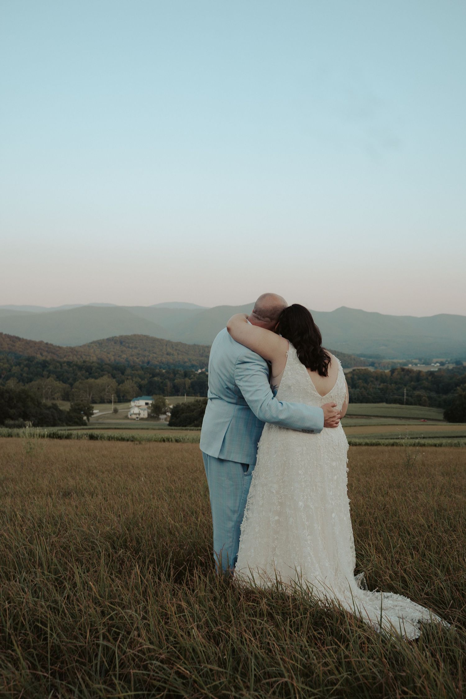 Groom in a light blue suit and bride hug each other facing away from the camera overlooking the Virginia Blue Ridge Mountain skyline