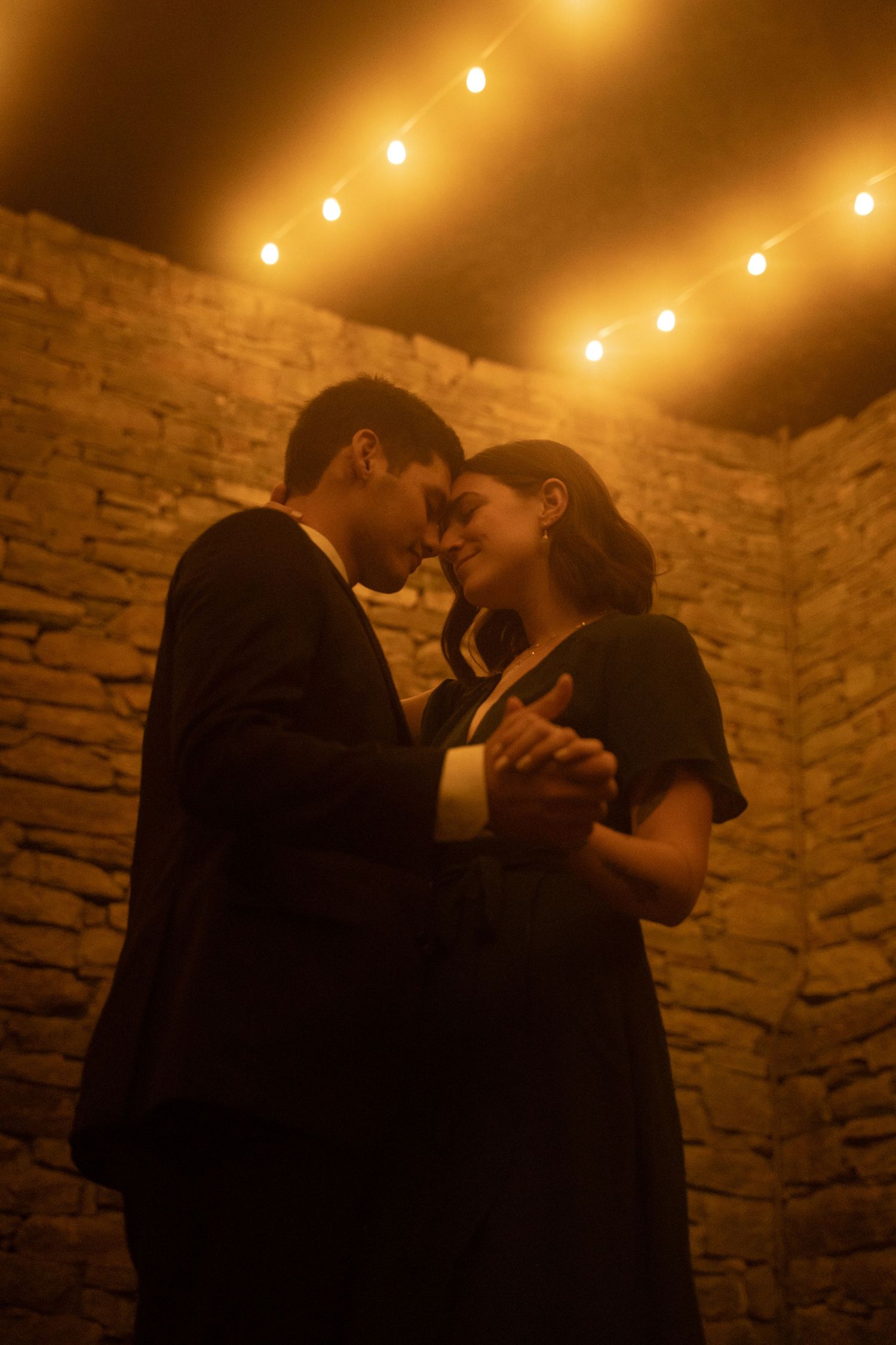Two wedding guests clasp hands and dance close under market lights in the ruins of the mill at Mill at Fine Creek outside Richmond Virginia