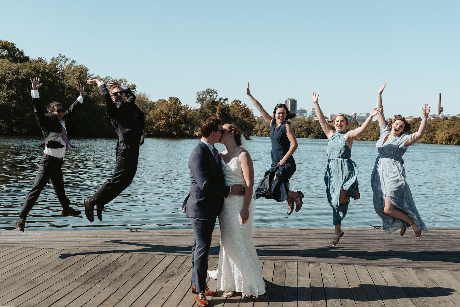 The wedding party jumps on a pier behind the newlyweds kissing before their ceremony at The Boathouse Rockett's Landing in Richmond Virginia