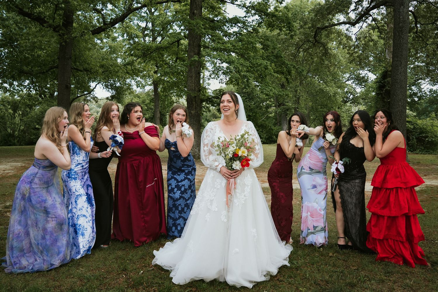 Bride in classic lace fall wedding photo holding jewel tone bouquet smiles while bridesmaids in rich red and blue gasp surrounding her
