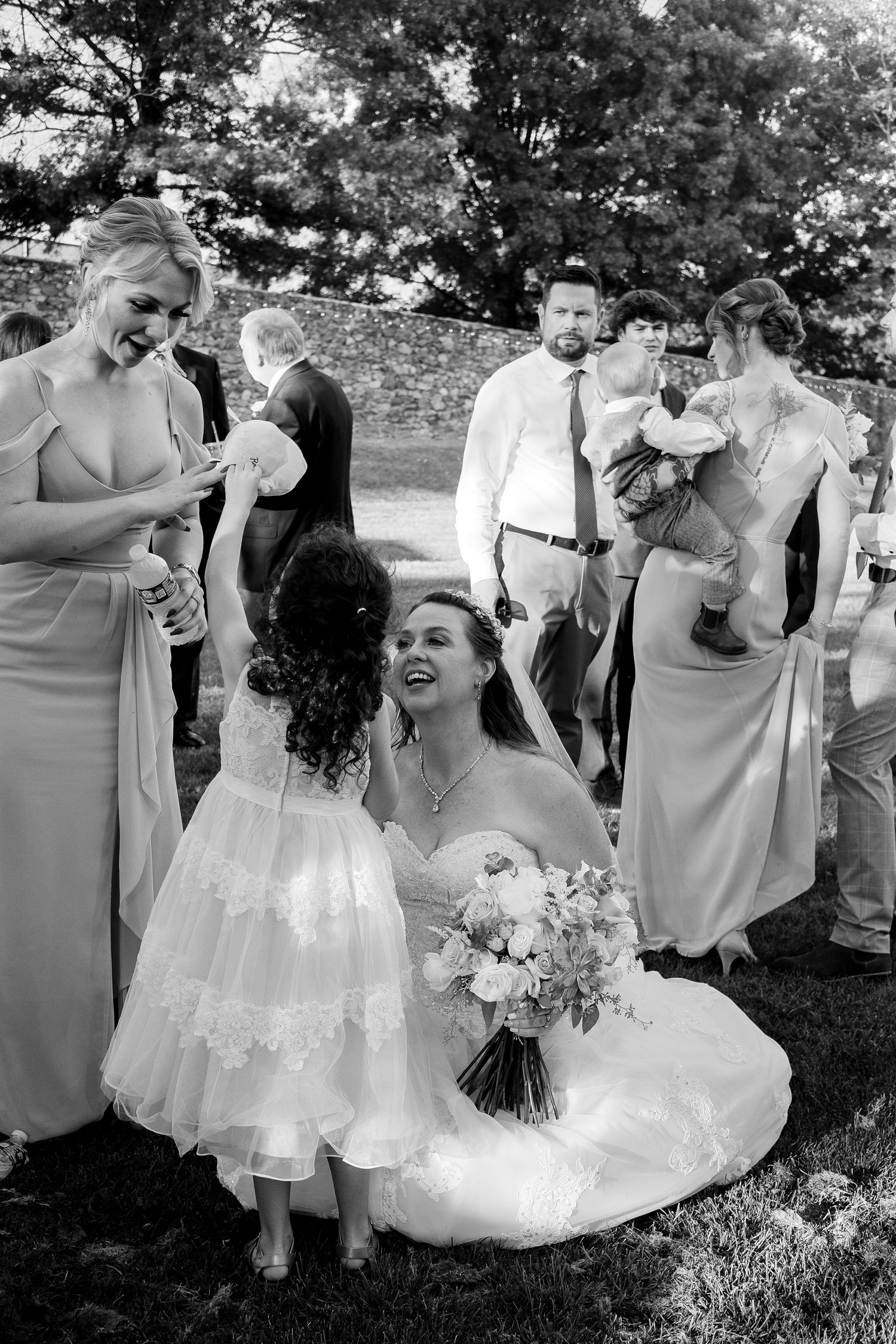 Bride in timeless wedding dress crouches to greet her granddaughter in classic black and white documentary style photograph
