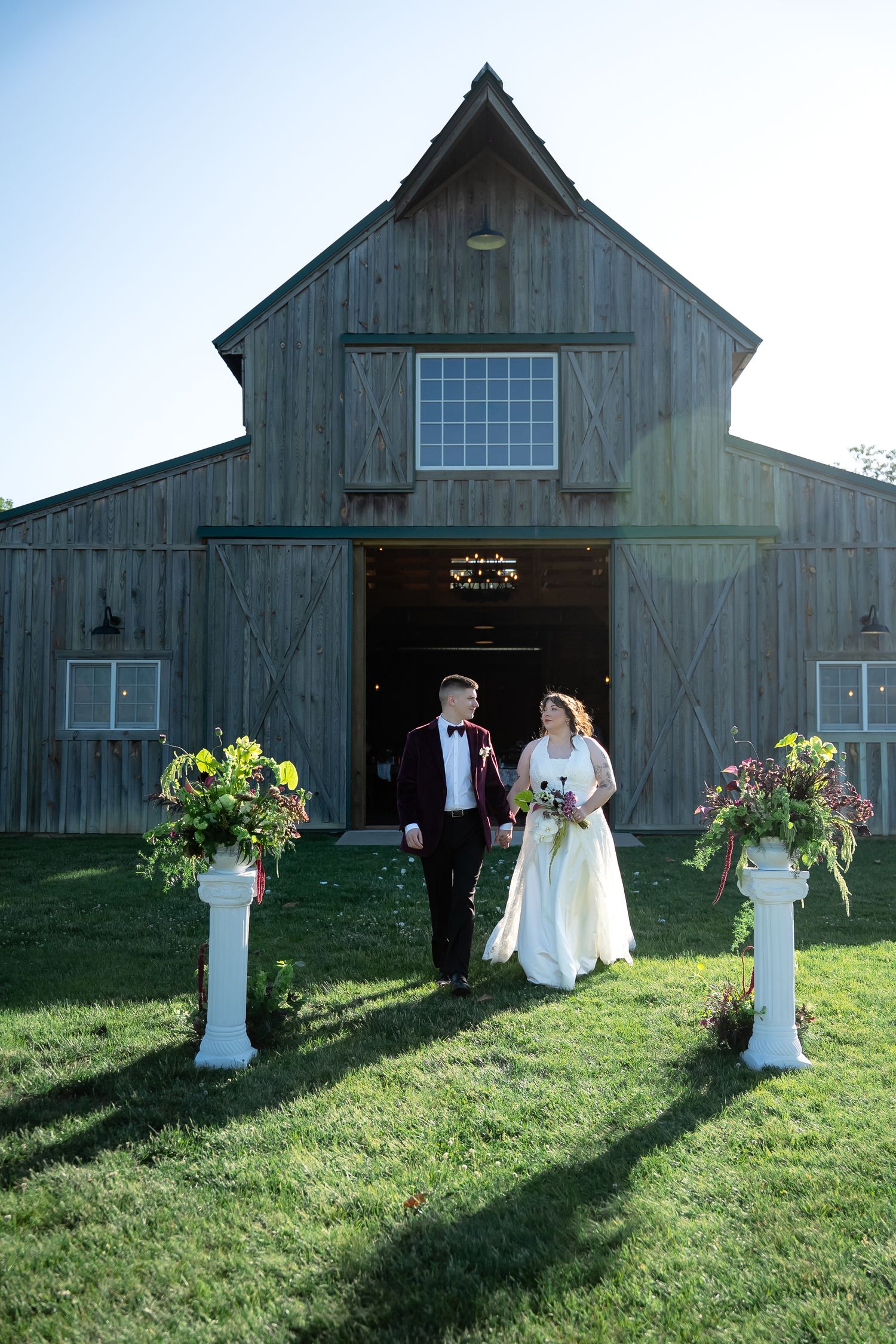 A bride and groom exit their wedding barn between two bouquets on pedestals while the low sun casts long shadows towards the caemra