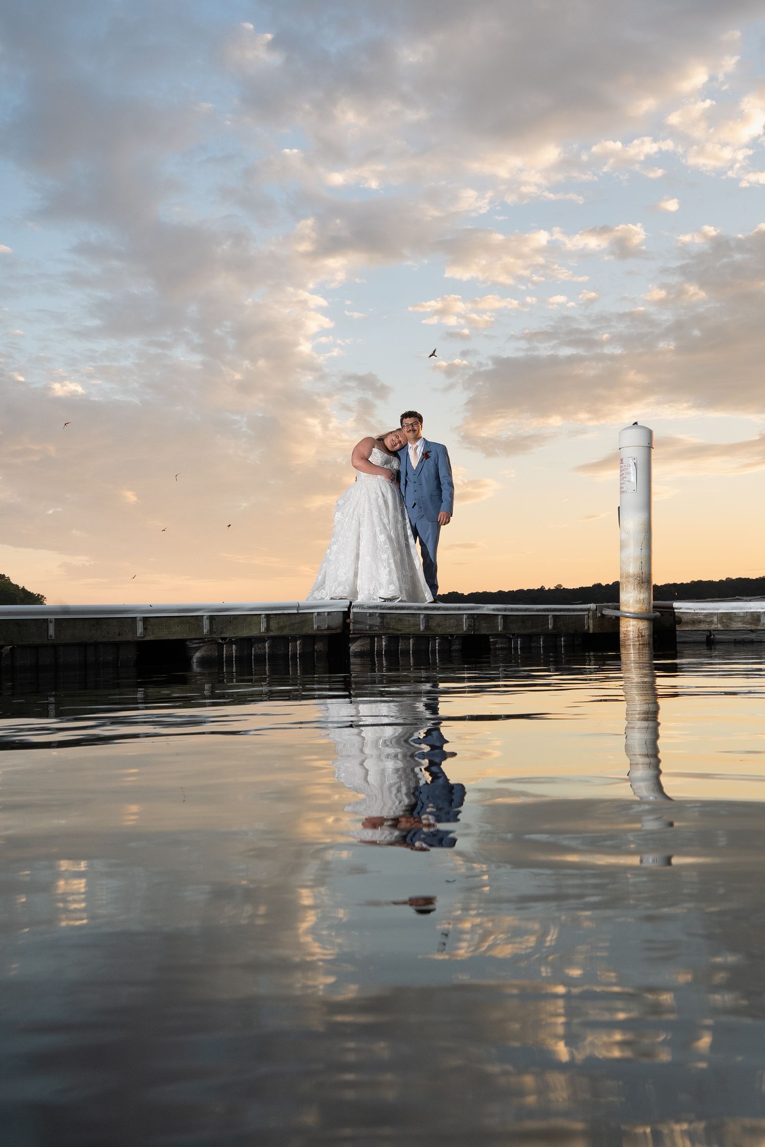 A bride and groom are reflected in a lake in the foreground at the Boatyard in Sunday Park in a modern yet classic natural wedding aesthetic