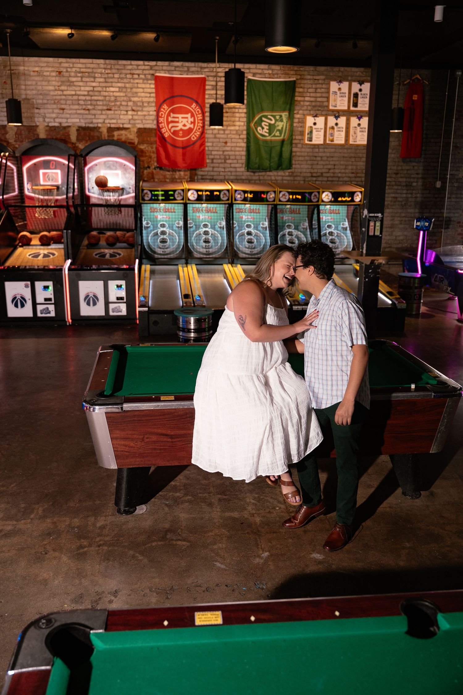 Cool girl engagement session at a bar, couple sits on pool table and kisses with arcade games in the background of this photo session