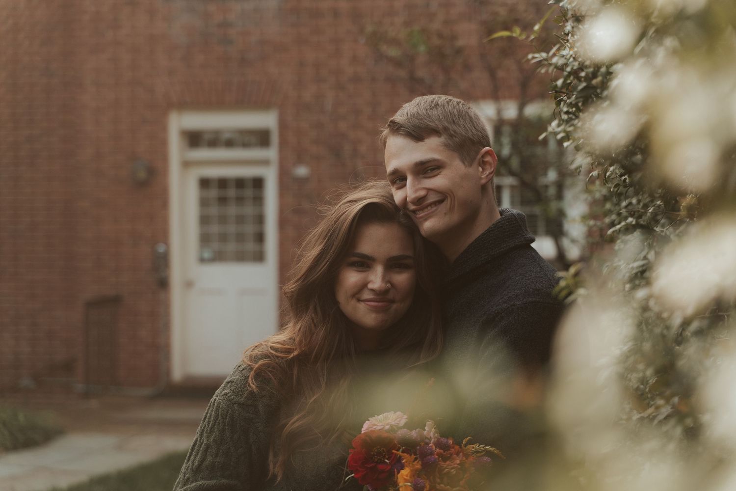 Fall engagement session aesthetic couple smiles at camera hugging with leaves in the foreground rich fall colored bouquet in woman's hands
