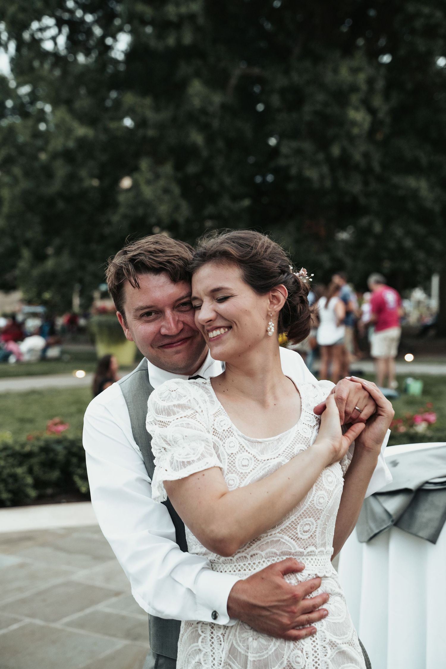 A groom embraces his bride from behind as she spins into his arms, as crowds in the background enjoy a summer evening at a museum courtyard