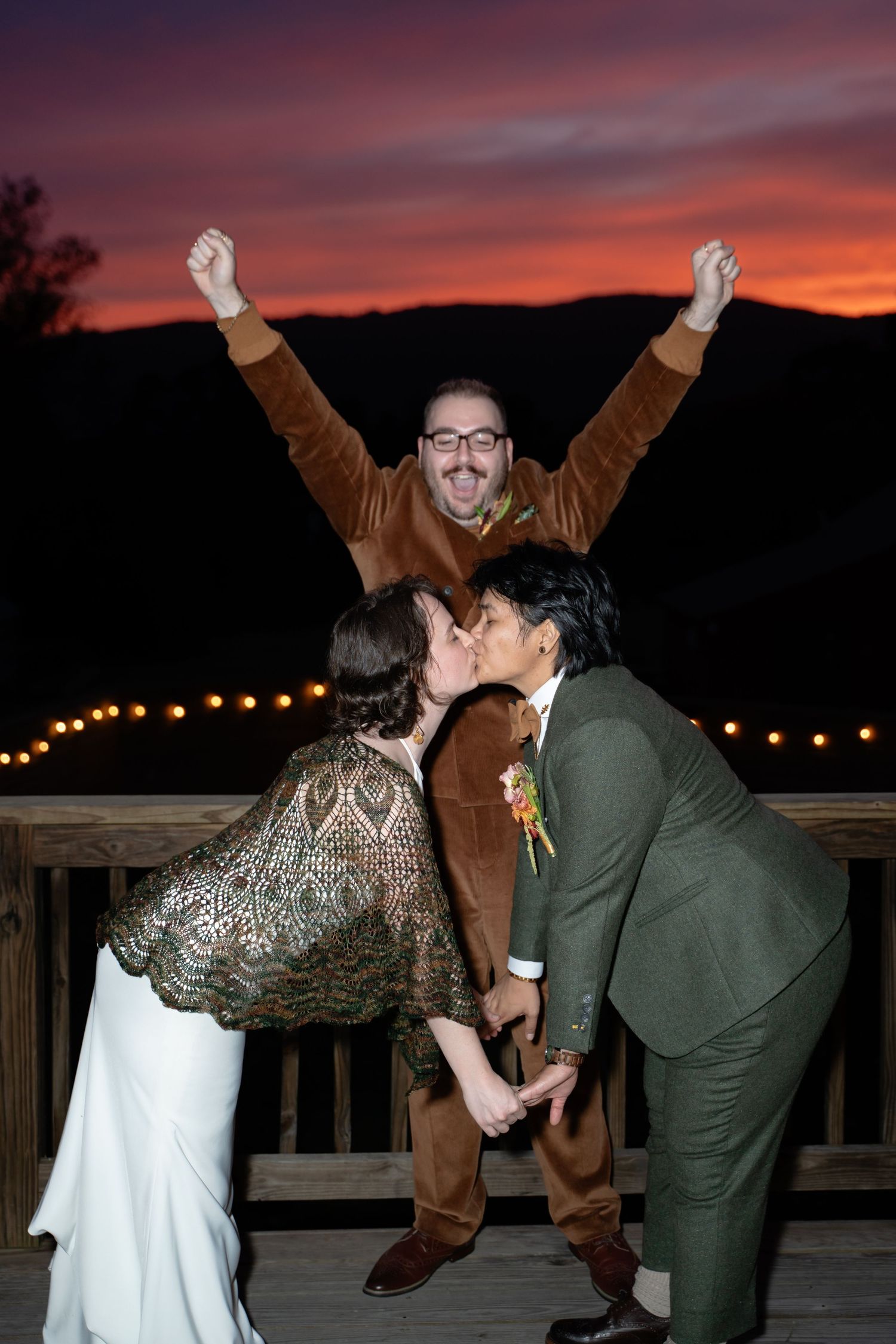 Wedding officiant celebrates behind two queer brides as they reenact their ceremonial kiss during cocktail hour in front of a sunset