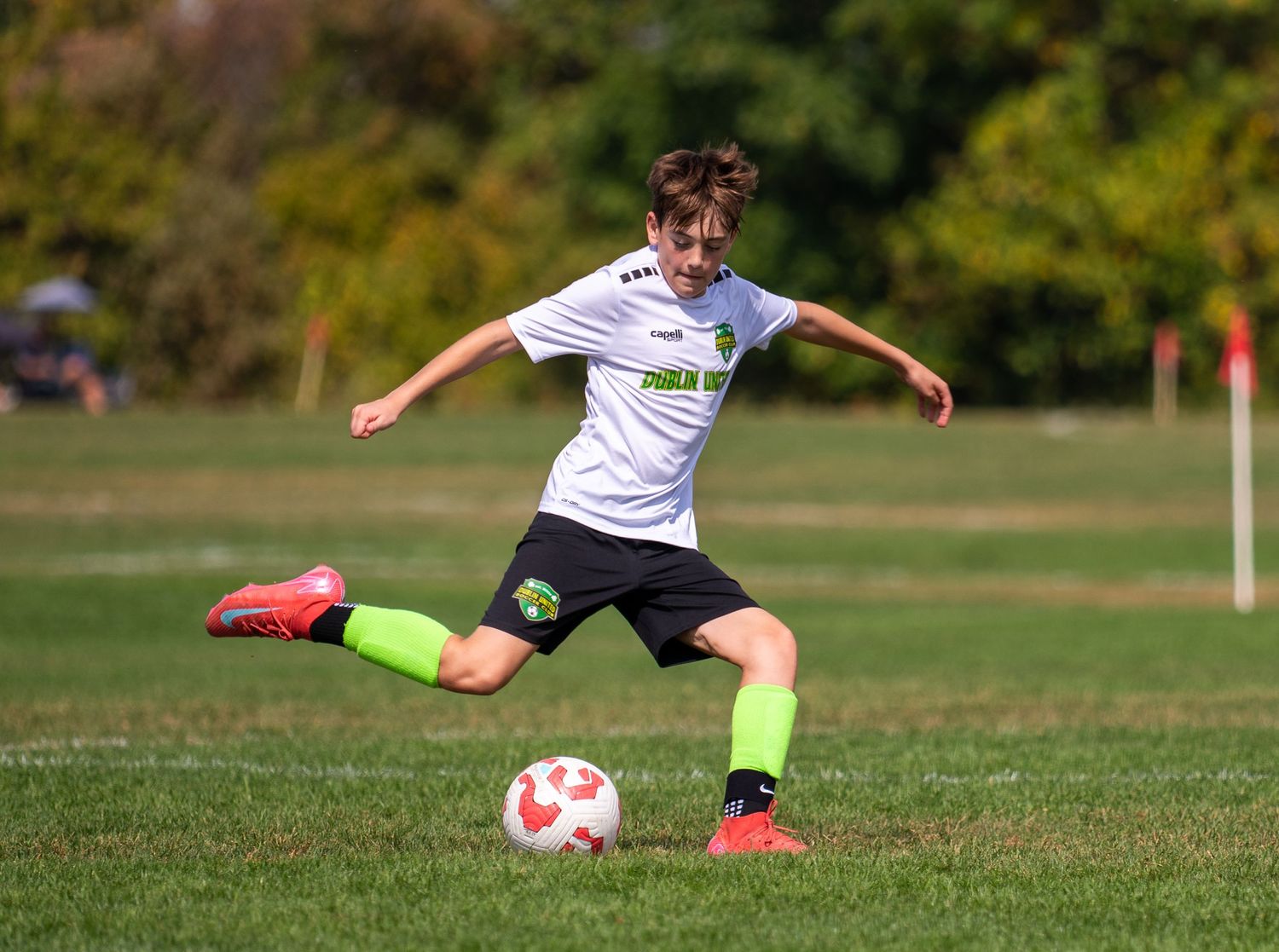 Youth soccer player in white and green uniform kicking a ball during an outdoor soccer game.