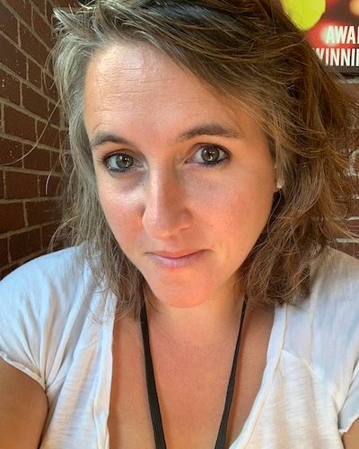 Woman with shoulder-length blonde hair wearing a white t-shirt and lanyard, taking a selfie in front of a brick wall.