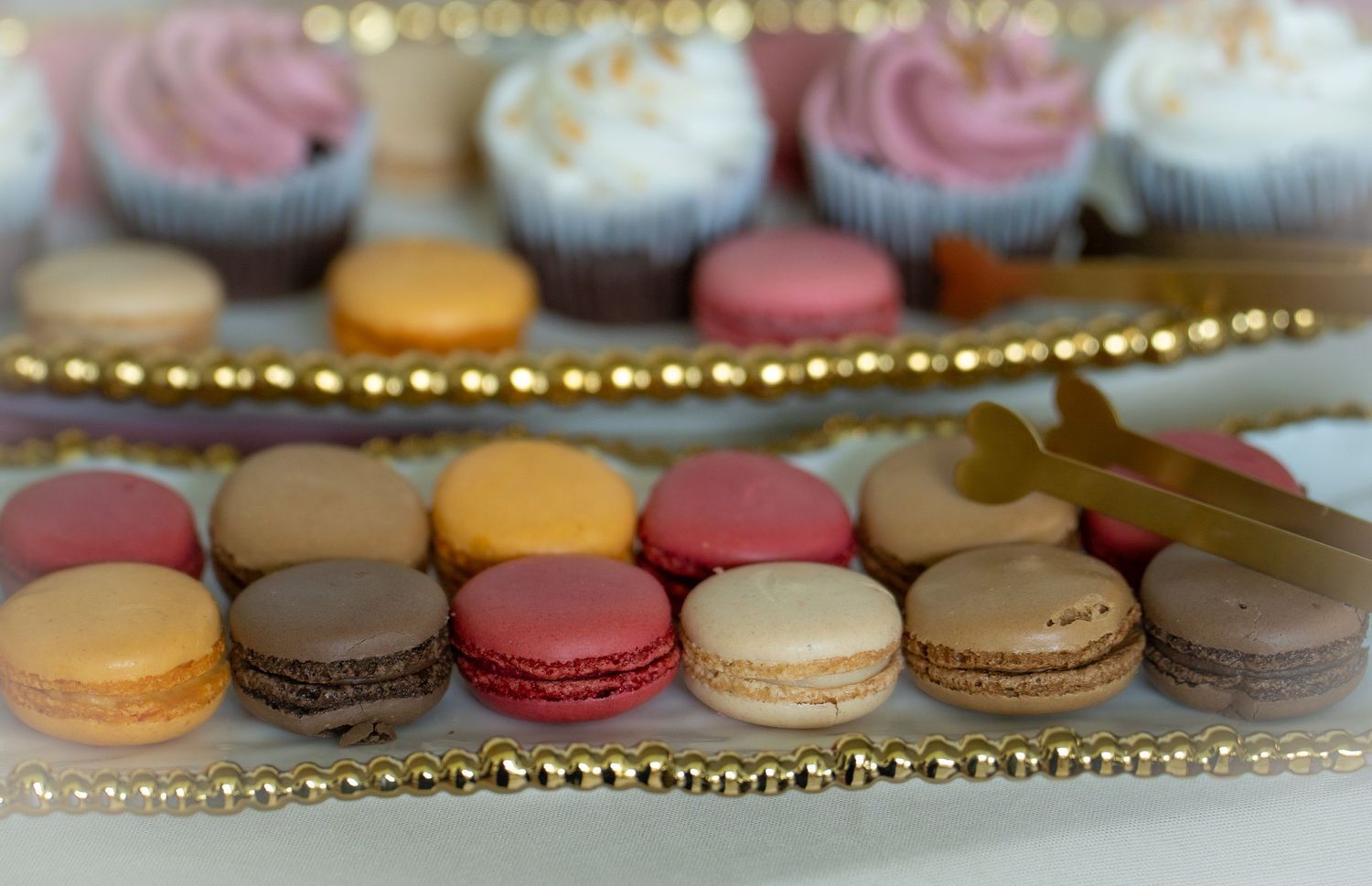 Assorted colorful French macarons and cupcakes displayed on gold-trimmed dessert table at wedding.