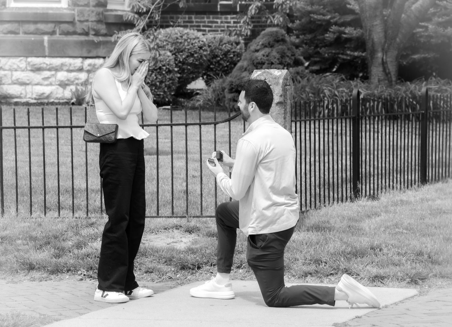 Man proposes on one knee to surprised woman outdoors near iron fence in black and white engagement photo.