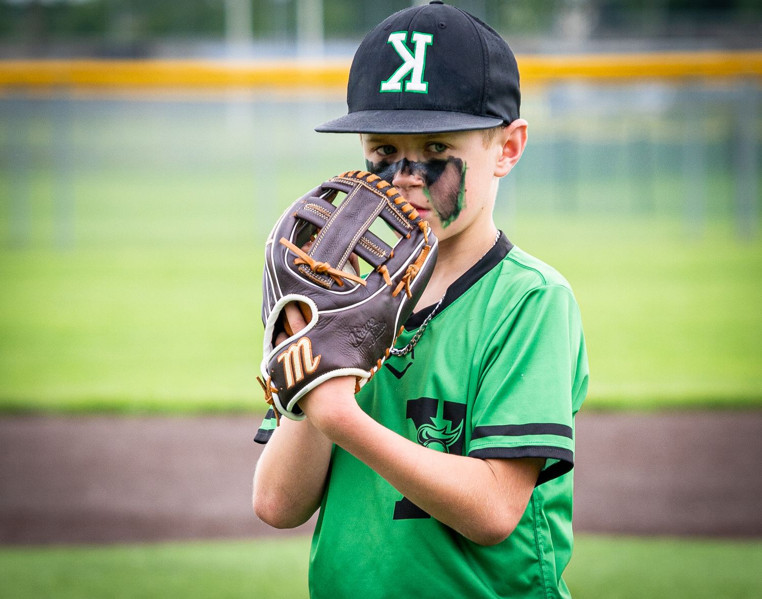 Youth baseball pitcher in green Knights jersey winds up with glove covering face on field.