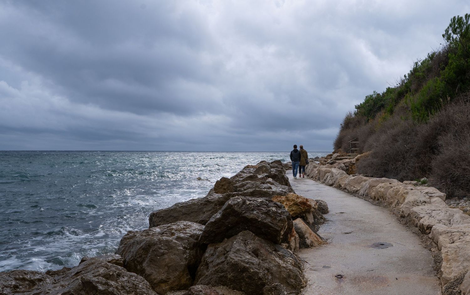 Two people walking along a rocky coastal path under dramatic cloudy skies.