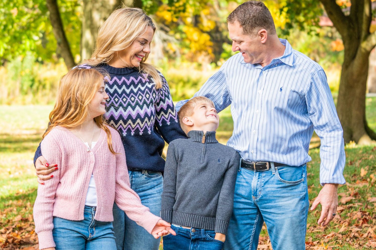 Family of four laughing together in autumn park, parents and two children holding hands among fall foliage.