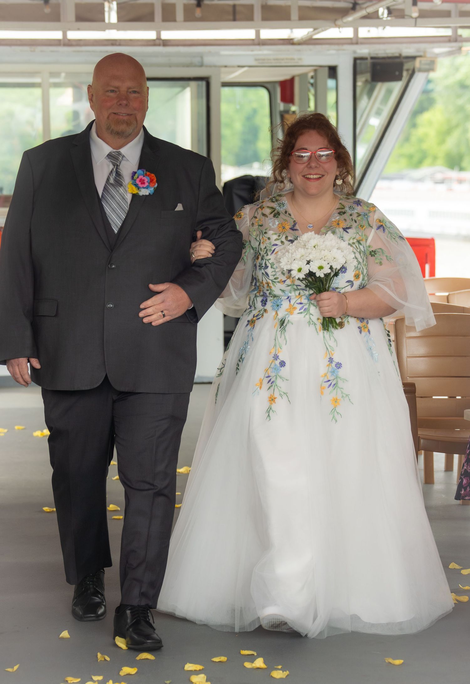 Bride in floral gown holding bouquet walks with groom in gray suit at wedding ceremony.