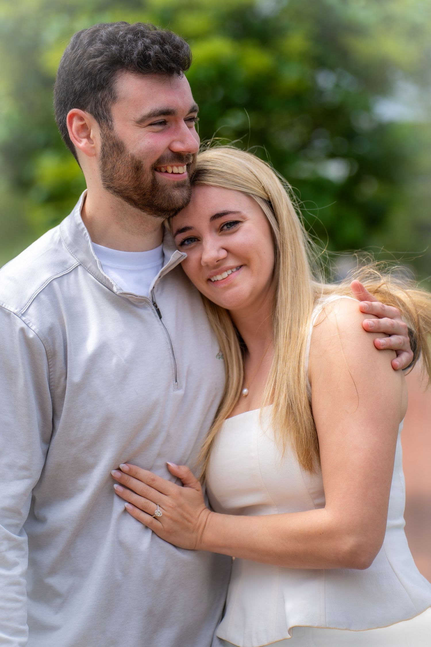 A smiling couple embracing outdoors with lush green trees in the background during an engagement shoot.