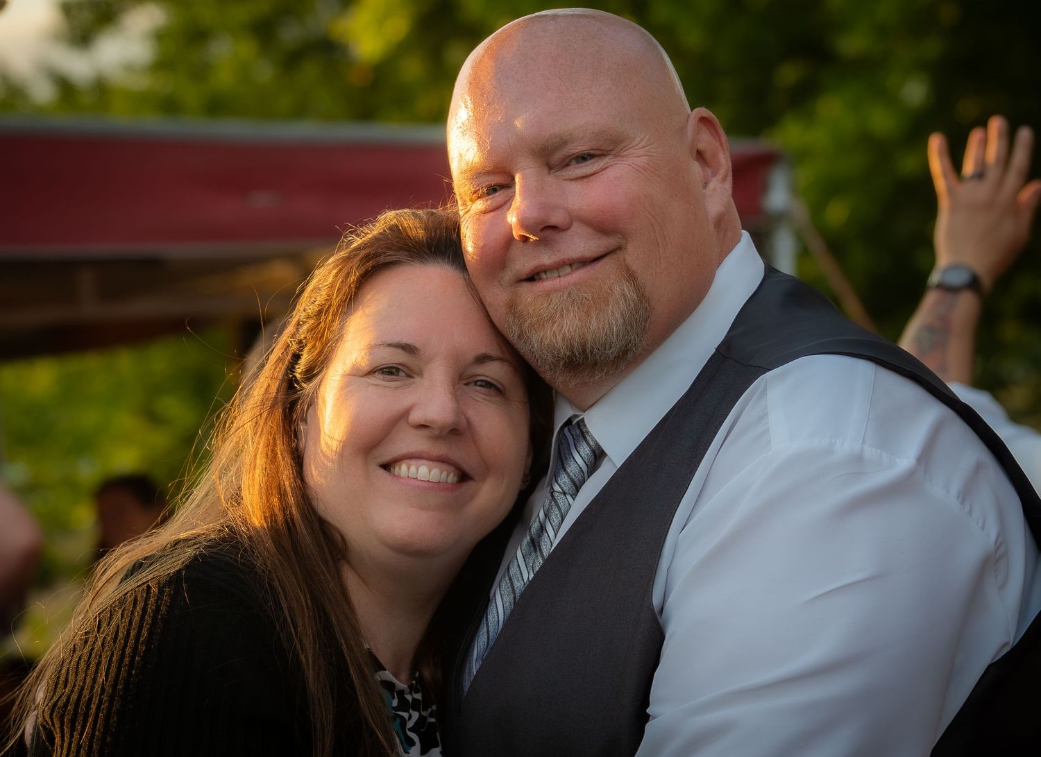 A smiling couple poses closely together outdoors at a casual evening event, man in vest and tie.