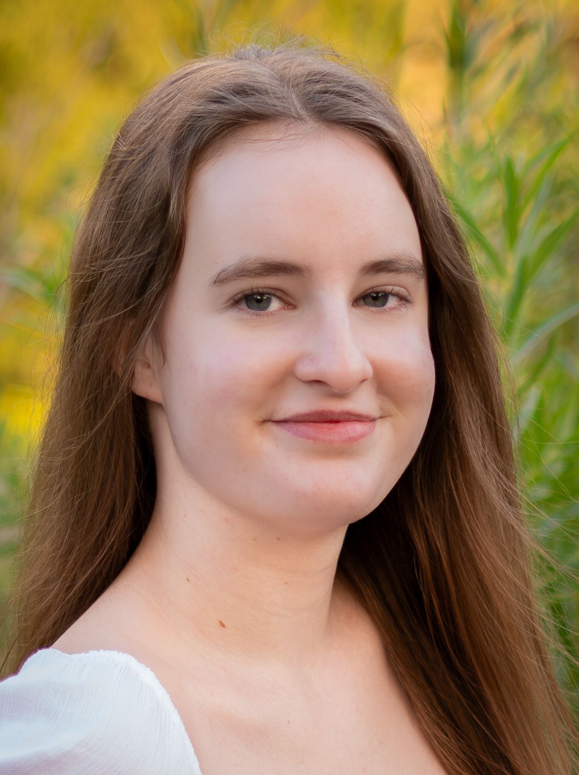 Young woman with long auburn hair smiling outdoors with green foliage background.