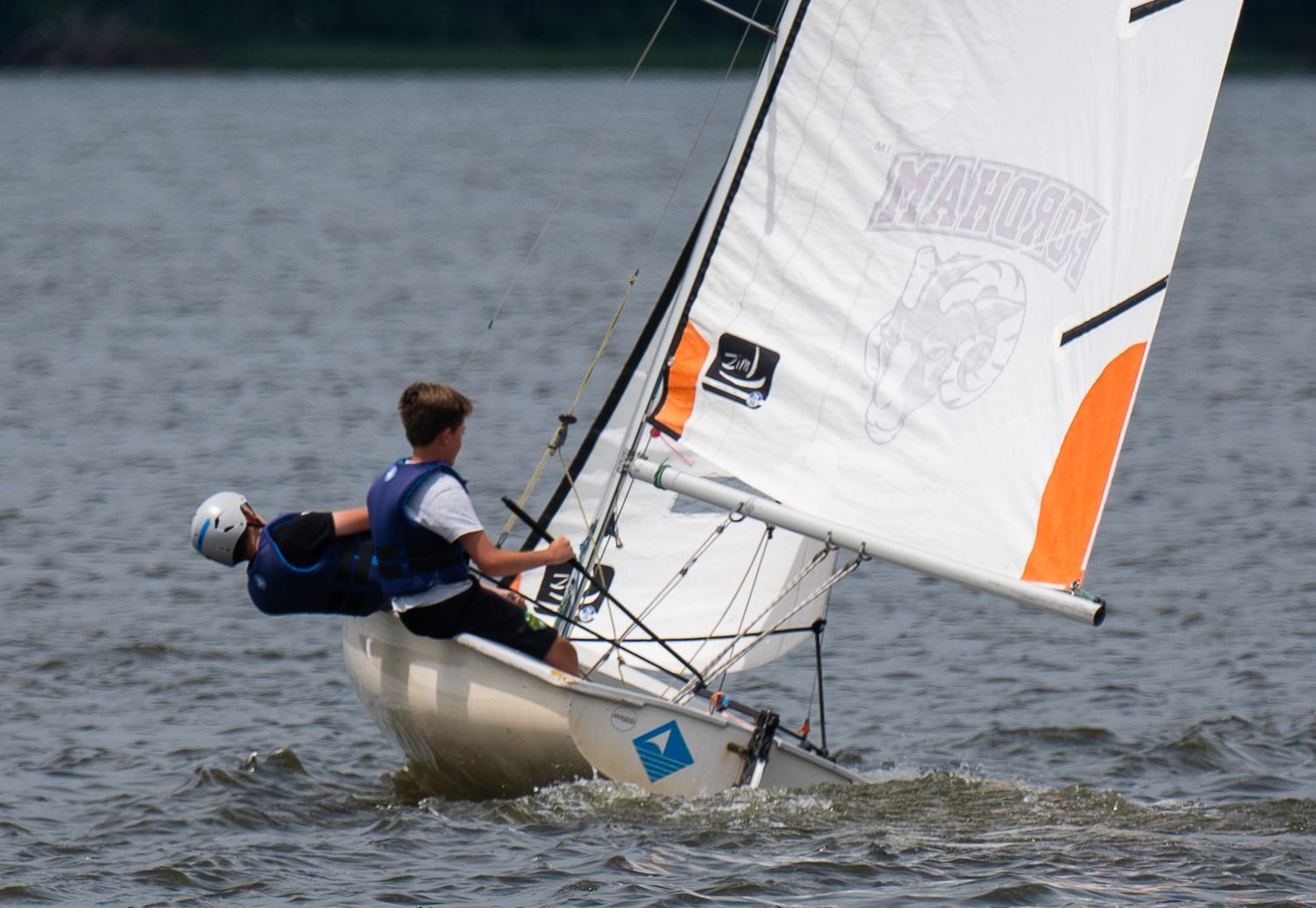 Two sailors navigate a small white and orange sailboat on calm lake waters during regatta.