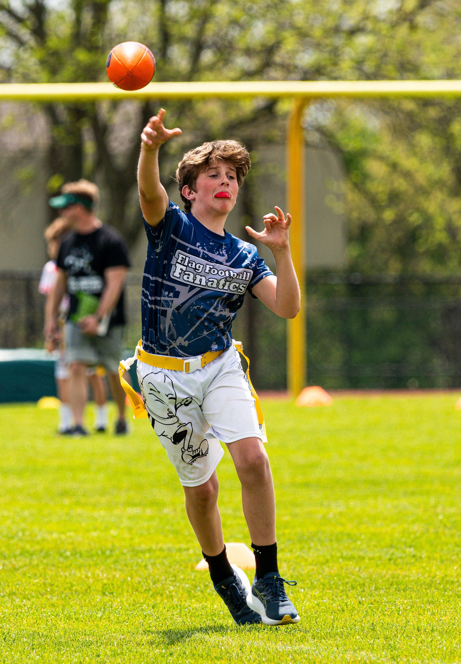 Young boy in blue jersey throwing a football during an outdoor flag football game on a sunny day.