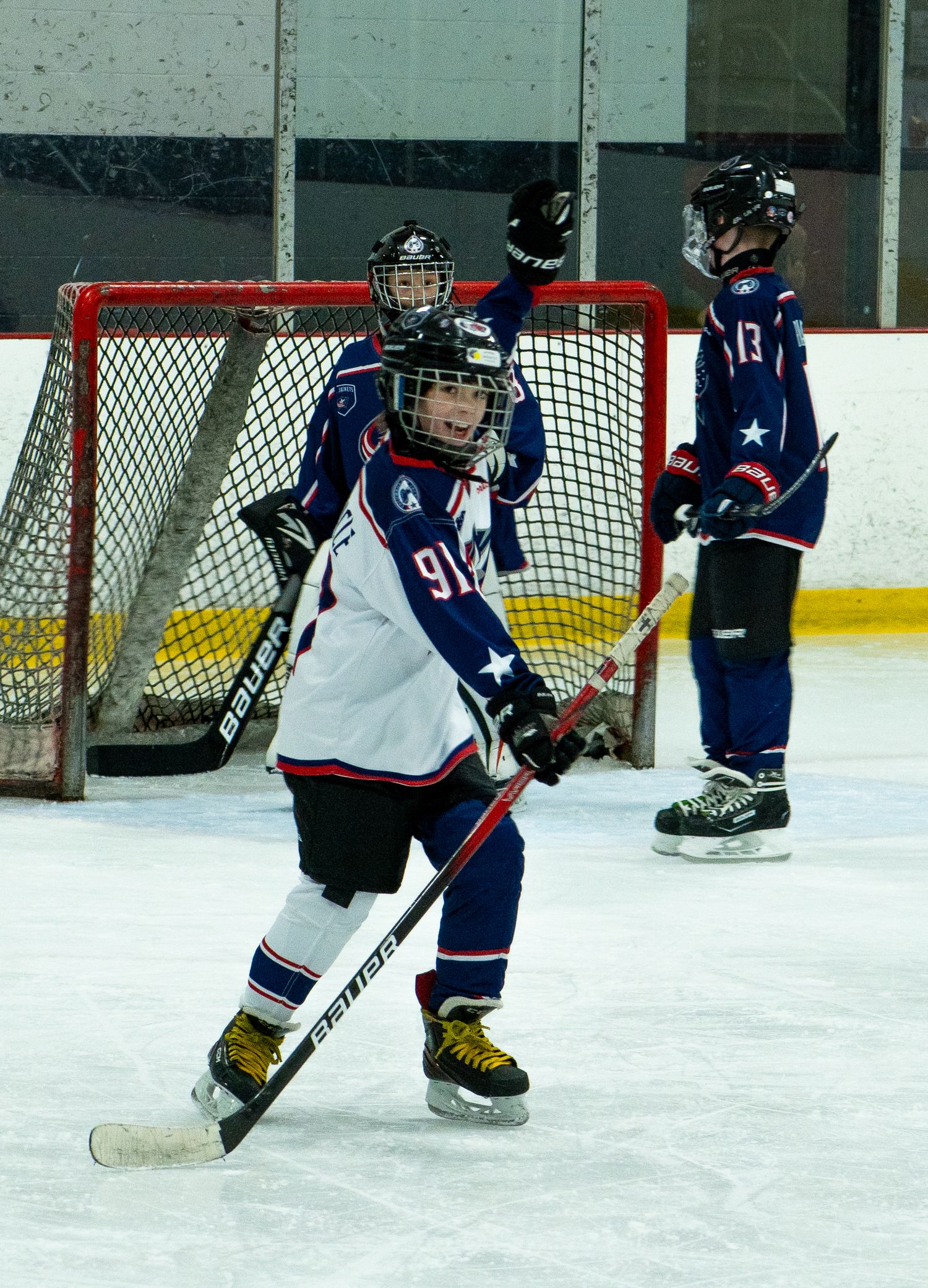 Youth hockey player celebrating goal in navy uniform near red net on indoor rink.
